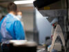 Lab scene with a technician in blue scrubs and mask at a workbench; foreground shows a medical device with red controls in focus elsewhere.