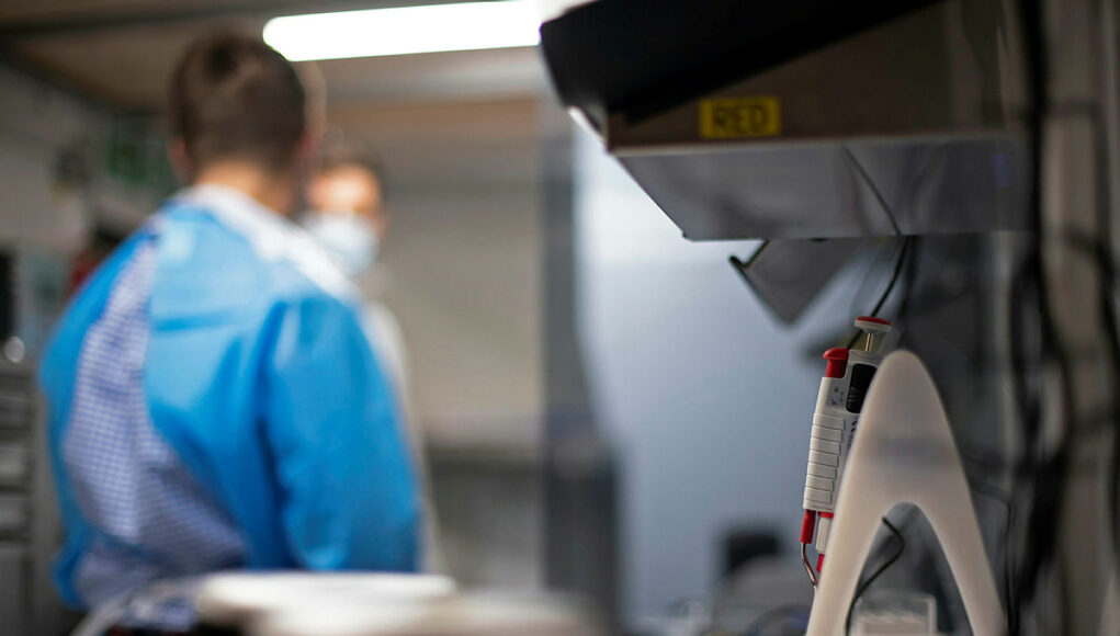 Lab scene with a technician in blue scrubs and mask at a workbench; foreground shows a medical device with red controls in focus elsewhere.
