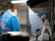 Lab scene with a technician in blue scrubs and mask at a workbench; foreground shows a medical device with red controls in focus elsewhere.