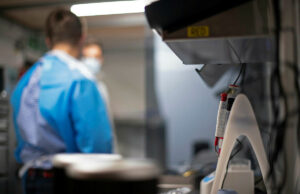 Lab scene with a technician in blue scrubs and mask at a workbench; foreground shows a medical device with red controls in focus elsewhere.