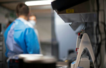 Lab scene with a technician in blue scrubs and mask at a workbench; foreground shows a medical device with red controls in focus elsewhere.