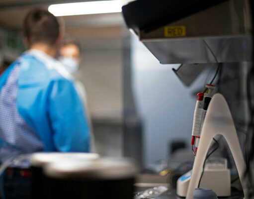 Lab scene with a technician in blue scrubs and mask at a workbench; foreground shows a medical device with red controls in focus elsewhere.