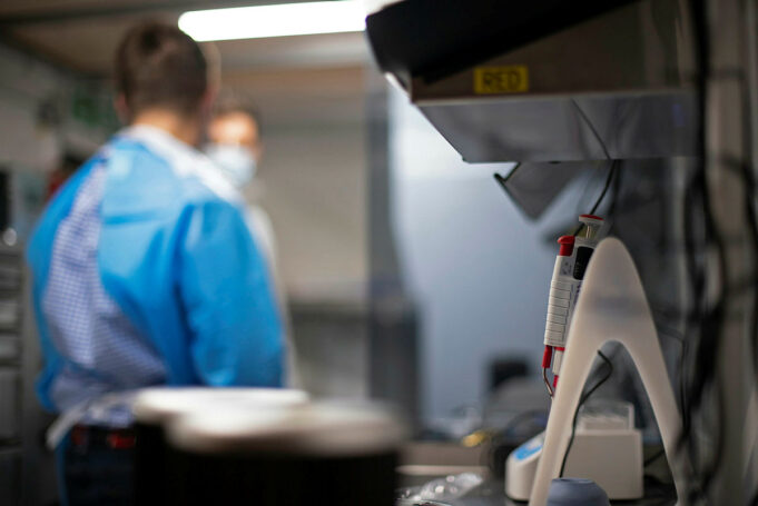 Lab scene with a technician in blue scrubs and mask at a workbench; foreground shows a medical device with red controls in focus elsewhere.