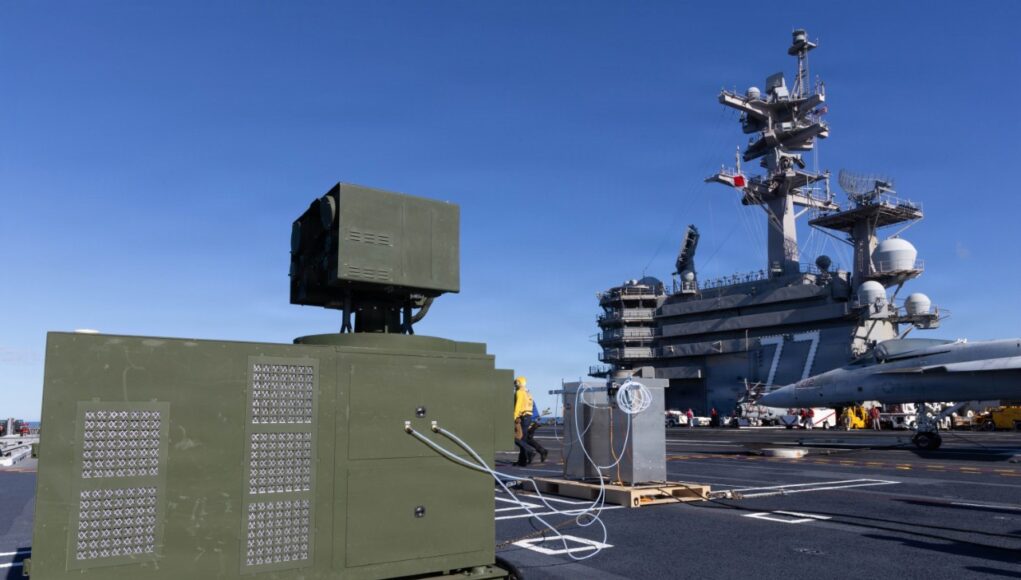 Green electrical equipment cabinet with vent panels on a naval carrier deck, cables connected, with a fighter jet in the background on the right.