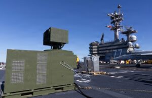 Green electrical equipment cabinet with vent panels on a naval carrier deck, cables connected, with a fighter jet in the background on the right.