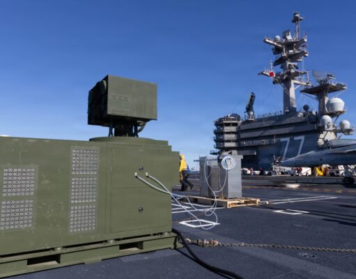 Green electrical equipment cabinet with vent panels on a naval carrier deck, cables connected, with a fighter jet in the background on the right.