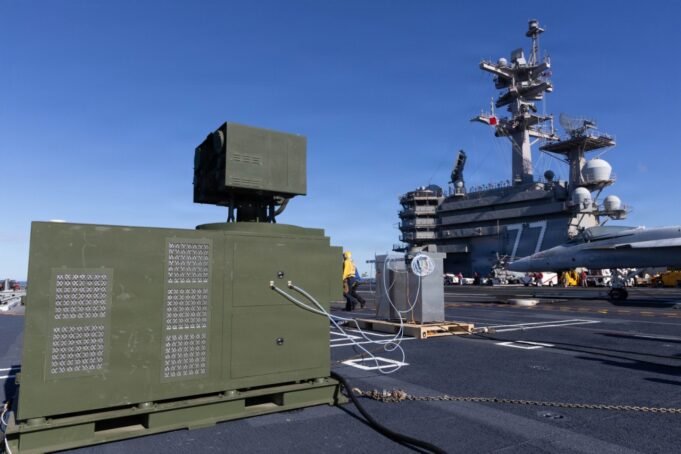 Green electrical equipment cabinet with vent panels on a naval carrier deck, cables connected, with a fighter jet in the background on the right.