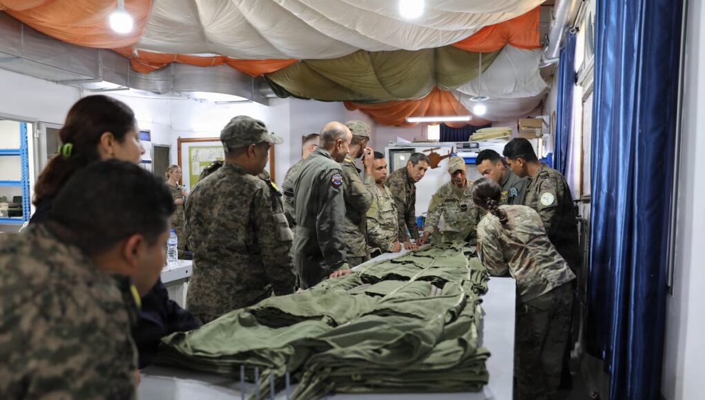 Group of soldiers in camouflage uniforms standing around a long table, inspecting and folding large stacks of green fabric gear inside a makeshift tent.