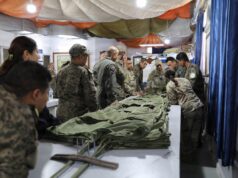 Group of soldiers in camouflage uniforms standing around a long table, inspecting and folding large stacks of green fabric gear inside a makeshift tent.