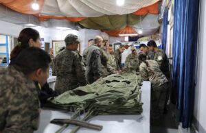 Group of soldiers in camouflage uniforms standing around a long table, inspecting and folding large stacks of green fabric gear inside a makeshift tent.