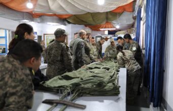 Group of soldiers in camouflage uniforms standing around a long table, inspecting and folding large stacks of green fabric gear inside a makeshift tent.