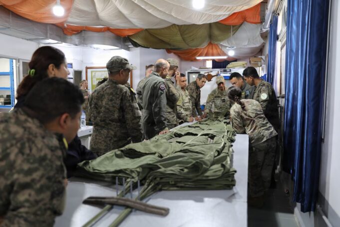 Group of soldiers in camouflage uniforms standing around a long table, inspecting and folding large stacks of green fabric gear inside a makeshift tent.