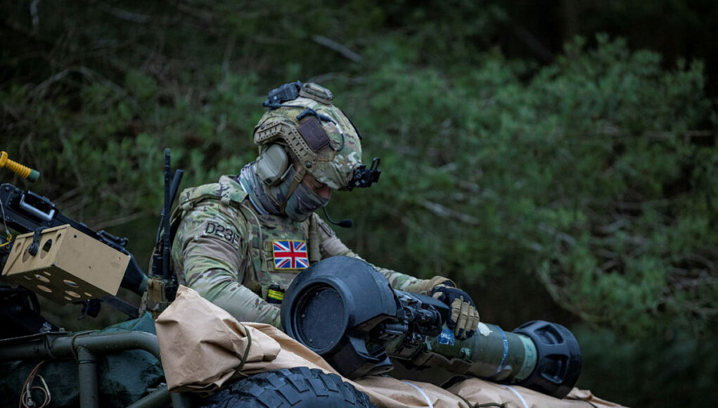 British soldier in full camouflage works on a large launcher mounted on a military vehicle in a wooded setting.