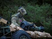 British soldier in full camouflage works on a large launcher mounted on a military vehicle in a wooded setting.