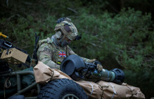 British soldier in full camouflage works on a large launcher mounted on a military vehicle in a wooded setting.