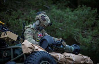 British soldier in full camouflage works on a large launcher mounted on a military vehicle in a wooded setting.