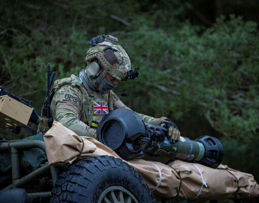 British soldier in full camouflage works on a large launcher mounted on a military vehicle in a wooded setting.