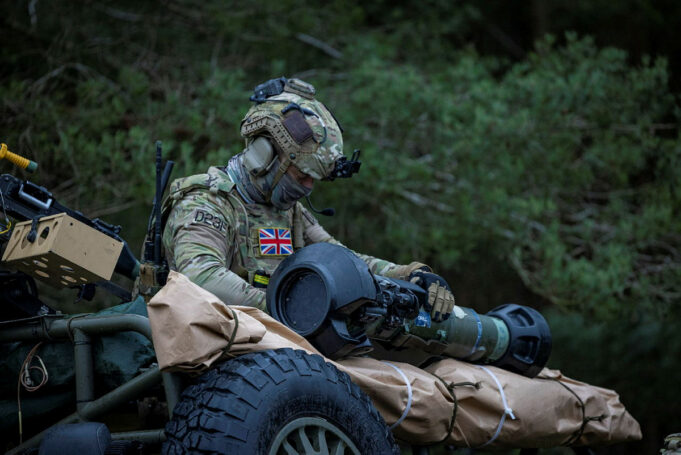 British soldier in full camouflage works on a large launcher mounted on a military vehicle in a wooded setting.