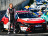 Race car driver in a black-and-white racing suit stands beside a red, white and black Toyota Corolla race car in the pit lane.