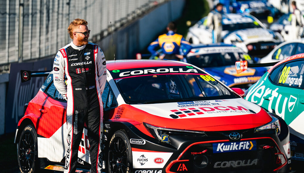 Race car driver in a black-and-white racing suit stands beside a red, white and black Toyota Corolla race car in the pit lane.