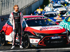 Race car driver in a black-and-white racing suit stands beside a red, white and black Toyota Corolla race car in the pit lane.