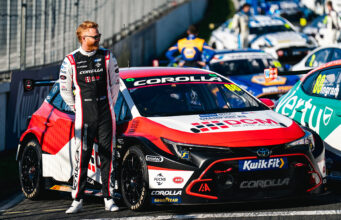 Race car driver in a black-and-white racing suit stands beside a red, white and black Toyota Corolla race car in the pit lane.