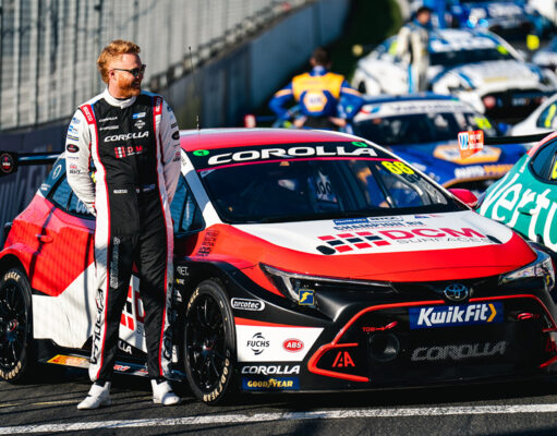Race car driver in a black-and-white racing suit stands beside a red, white and black Toyota Corolla race car in the pit lane.