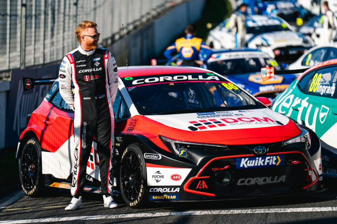 Race car driver in a black-and-white racing suit stands beside a red, white and black Toyota Corolla race car in the pit lane.