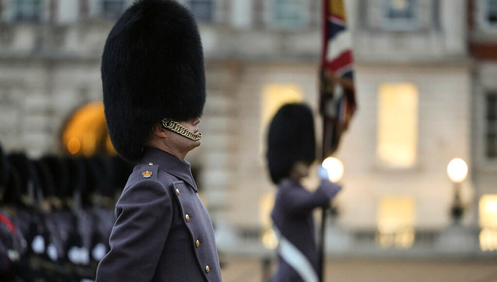 Beefeater guard in a tall fur hat stands in profile during a ceremonial parade, with another guard and a flag in the blurred background