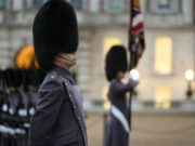 Beefeater guard in a tall fur hat stands in profile during a ceremonial parade, with another guard and a flag in the blurred background