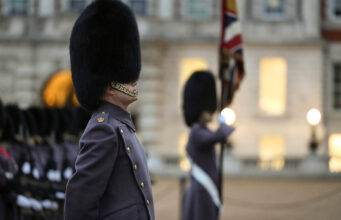 Beefeater guard in a tall fur hat stands in profile during a ceremonial parade, with another guard and a flag in the blurred background