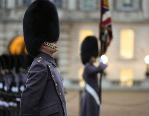 Beefeater guard in a tall fur hat stands in profile during a ceremonial parade, with another guard and a flag in the blurred background