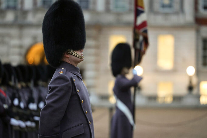 Beefeater guard in a tall fur hat stands in profile during a ceremonial parade, with another guard and a flag in the blurred background
