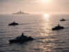 Hybrid ASW push raises command, comms and legal challenges Silhouetted naval ships sailing in calm ocean at sunset, with a large vessel visible on the horizon.