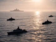 Silhouetted naval ships sailing in calm ocean at sunset, with a large vessel visible on the horizon.