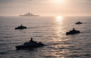 Silhouetted naval ships sailing in calm ocean at sunset, with a large vessel visible on the horizon.