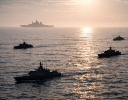 Silhouetted naval ships sailing in calm ocean at sunset, with a large vessel visible on the horizon.