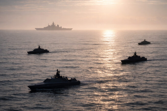 Silhouetted naval ships sailing in calm ocean at sunset, with a large vessel visible on the horizon.