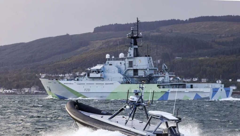 A large gray military ship with angular camouflage on the hull sails past while a fast inflatable boat speeds in choppy water in the foreground.