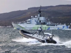 A large gray military ship with angular camouflage on the hull sails past while a fast inflatable boat speeds in choppy water in the foreground.