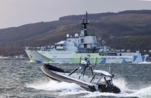 A large gray military ship with angular camouflage on the hull sails past while a fast inflatable boat speeds in choppy water in the foreground.