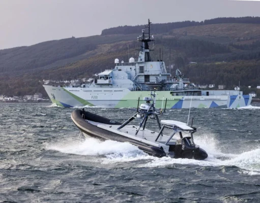 A large gray military ship with angular camouflage on the hull sails past while a fast inflatable boat speeds in choppy water in the foreground.