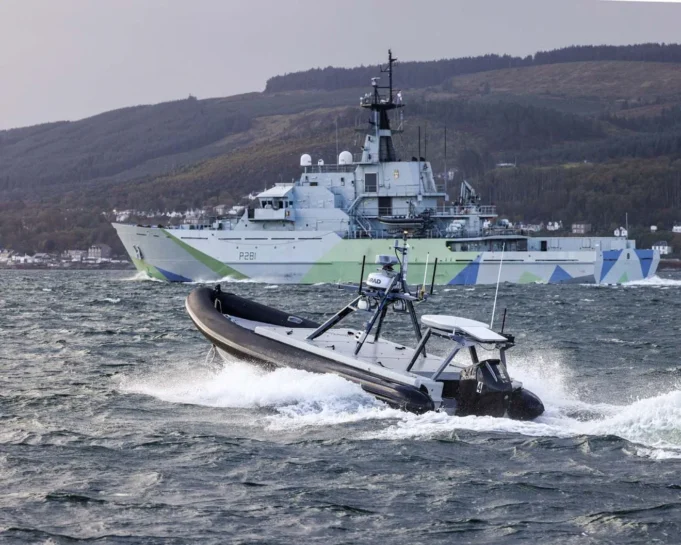 A large gray military ship with angular camouflage on the hull sails past while a fast inflatable boat speeds in choppy water in the foreground.