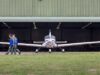 Small propeller plane facing viewer as two people in flight suits walk away with blue folders toward a hangar.