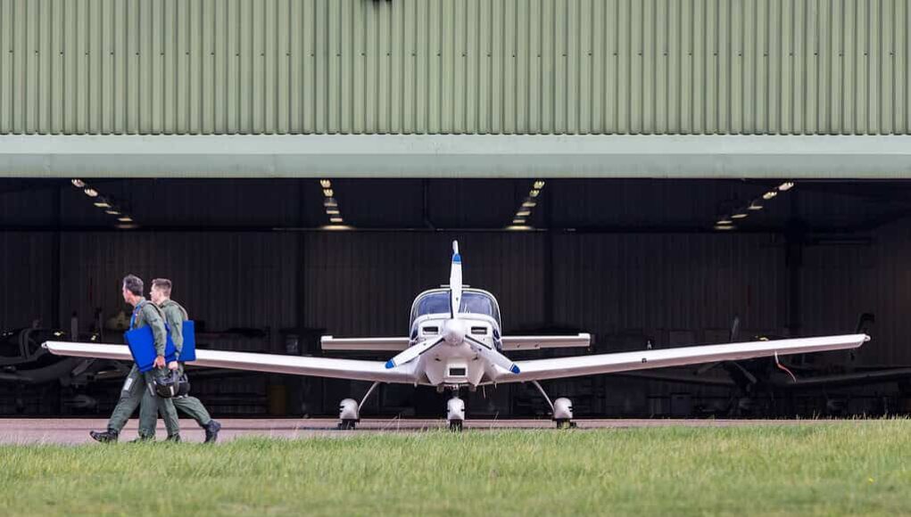 Small propeller plane facing viewer as two people in flight suits walk away with blue folders toward a hangar.