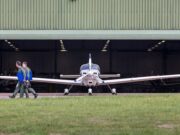 Small propeller plane facing viewer as two people in flight suits walk away with blue folders toward a hangar.