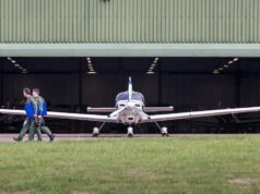 Small propeller plane facing viewer as two people in flight suits walk away with blue folders toward a hangar.