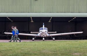 Small propeller plane facing viewer as two people in flight suits walk away with blue folders toward a hangar.