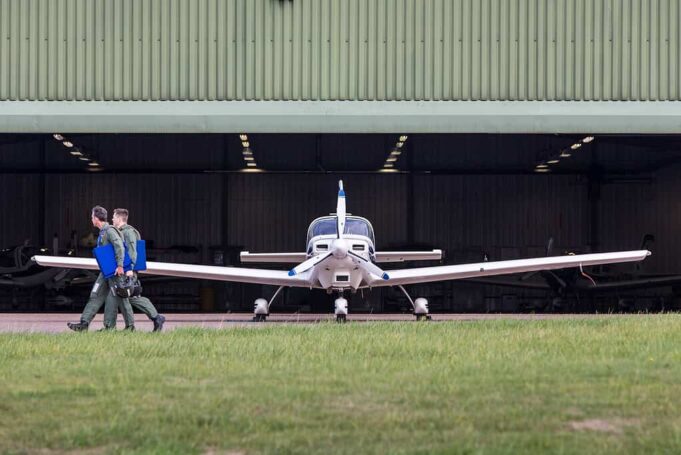 Small propeller plane facing viewer as two people in flight suits walk away with blue folders toward a hangar.