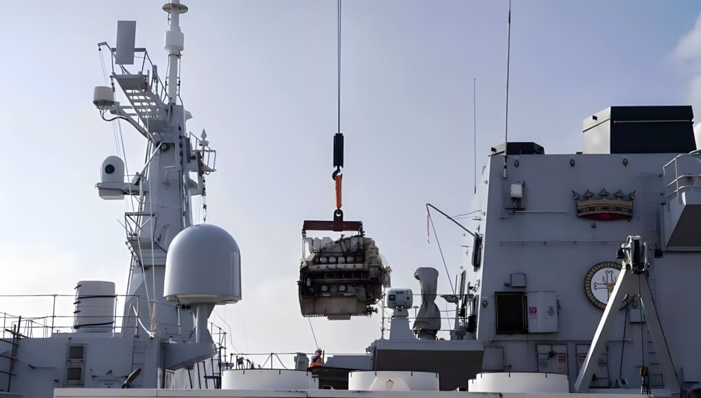 Crane on a gray warship hoists a heavy engine or equipment over the deck.